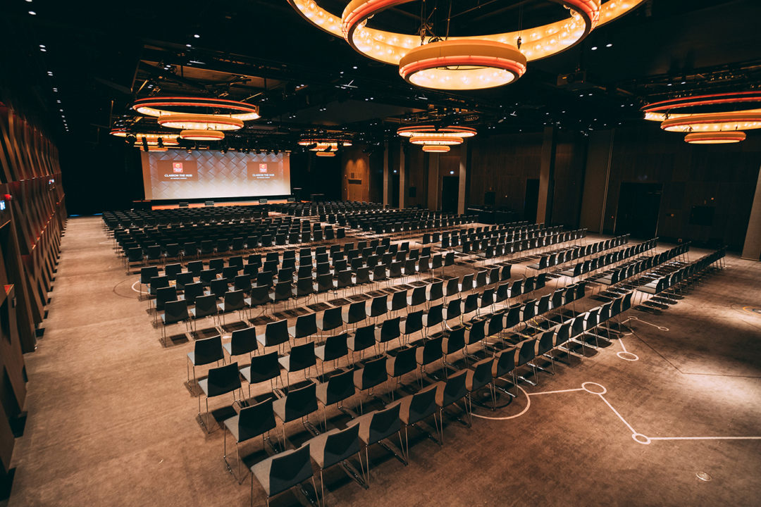 En stor og tom konferansesal med hundrevis av stoler. Storskjerm i front og moderne lamper i taket.
