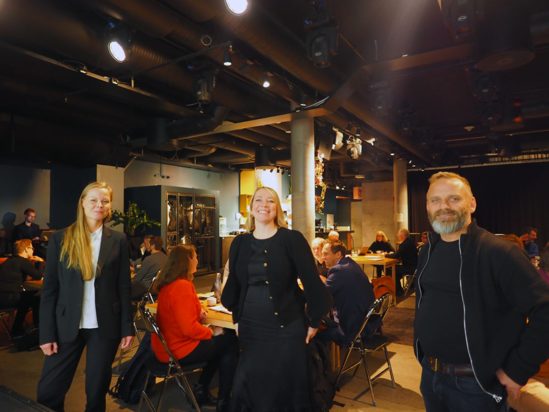 Emelie, Maren og Bjørnar står oppreist og smiler mot kamera. Bak dem sitter flere gruppe med mennesker og har workshop rundt flere bord.